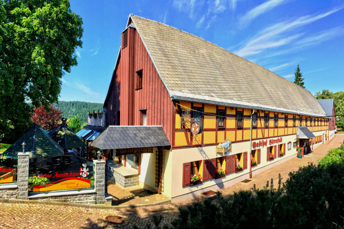 Ein großes, traditionelles Fachwerkgebäude mit roten und gelben Wänden, Blumenkästen und einem Schild mit der Aufschrift Gasthof Bärenfels unter blauem Himmel, umgeben von Grün und einem Kopfsteinpflasterweg, verkörpert den Charme eines klassischen Naturhotels.