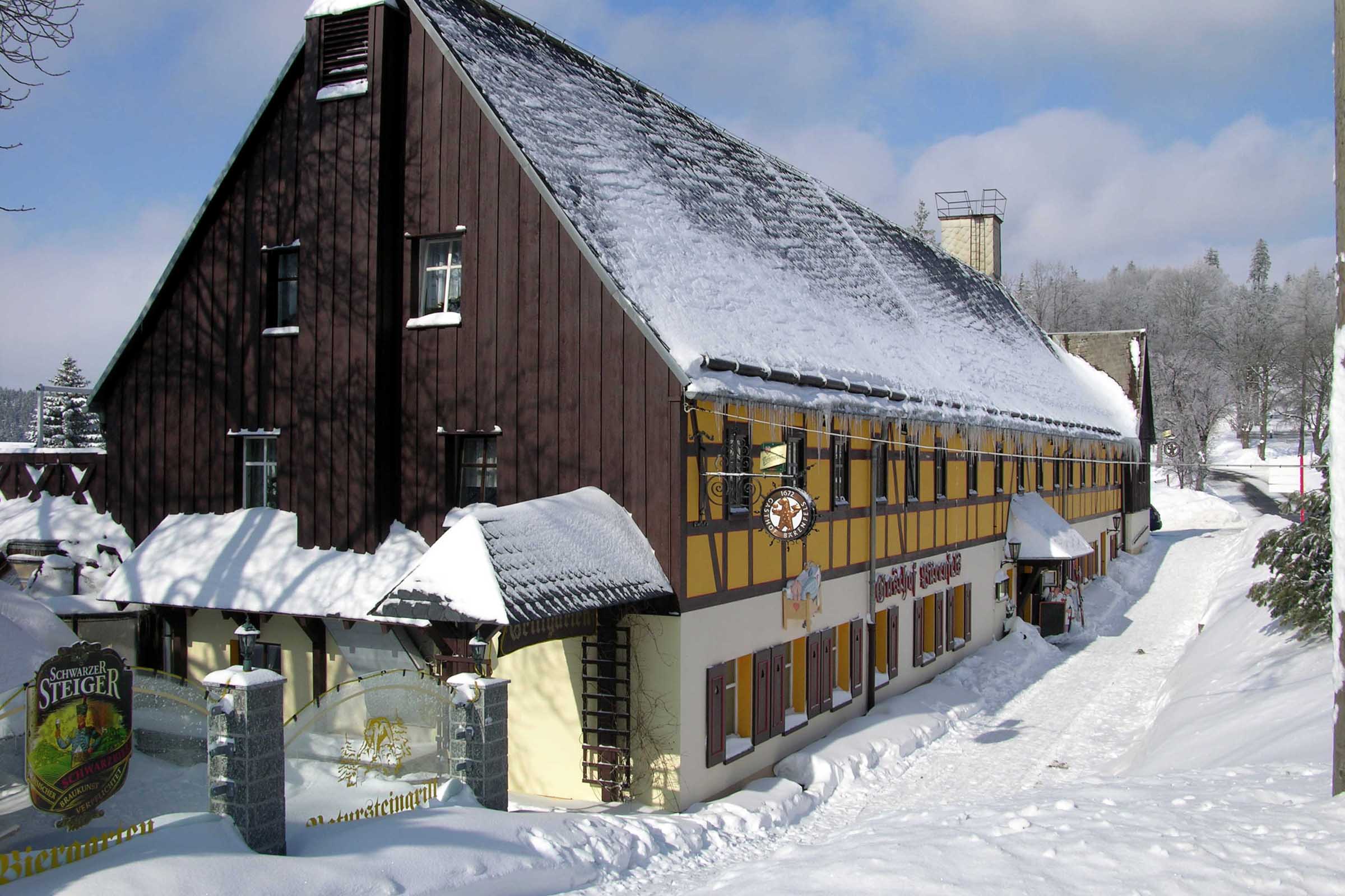 Das Naturhotel Gasthof Bärenfels, ein traditionelles Gebäude mit braunem und gelbem Fachwerk, ist mit Schnee bedeckt, umgeben von schneebedeckten Bäumen und einem strahlend blauen Himmel, der eine winterliche Szene in ländlicher Umgebung schafft.