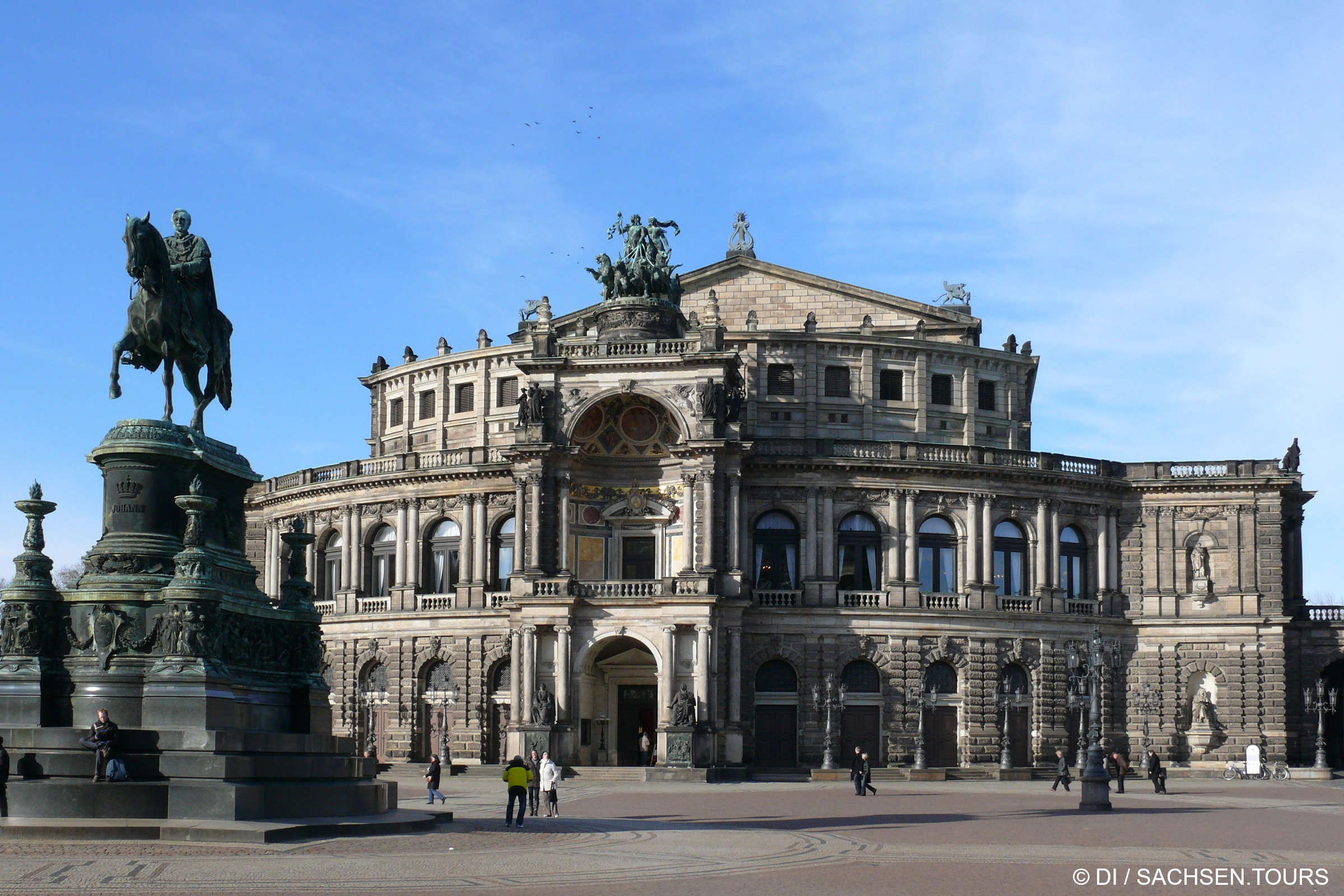 Die Semperoper Dresden ist ein großartiges historisches Opernhaus mit einer kunstvollen Architektur, großen Bogenfenstern und einem zentralen Eingang. Davor ziert ein bronzenes Reiterstandbild den offenen Dresdner Platz, auf dem man flanieren kann.