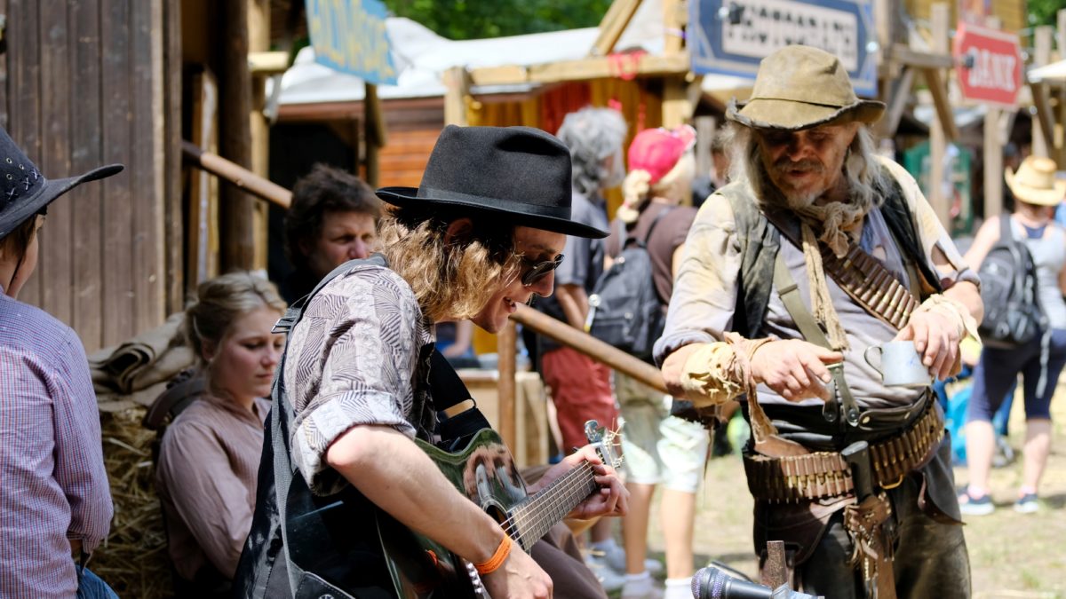 Ein Mann mit schwarzem Hut und Sonnenbrille spielt bei den Karl-May-Festtagen im Freien Gitarre, während ein anderer Mann, gekleidet wie ein Cowboy, zuschaut. Im Hintergrund stehen mehrere Personen in alter Westernkleidung bei dieser Themenveranstaltung.