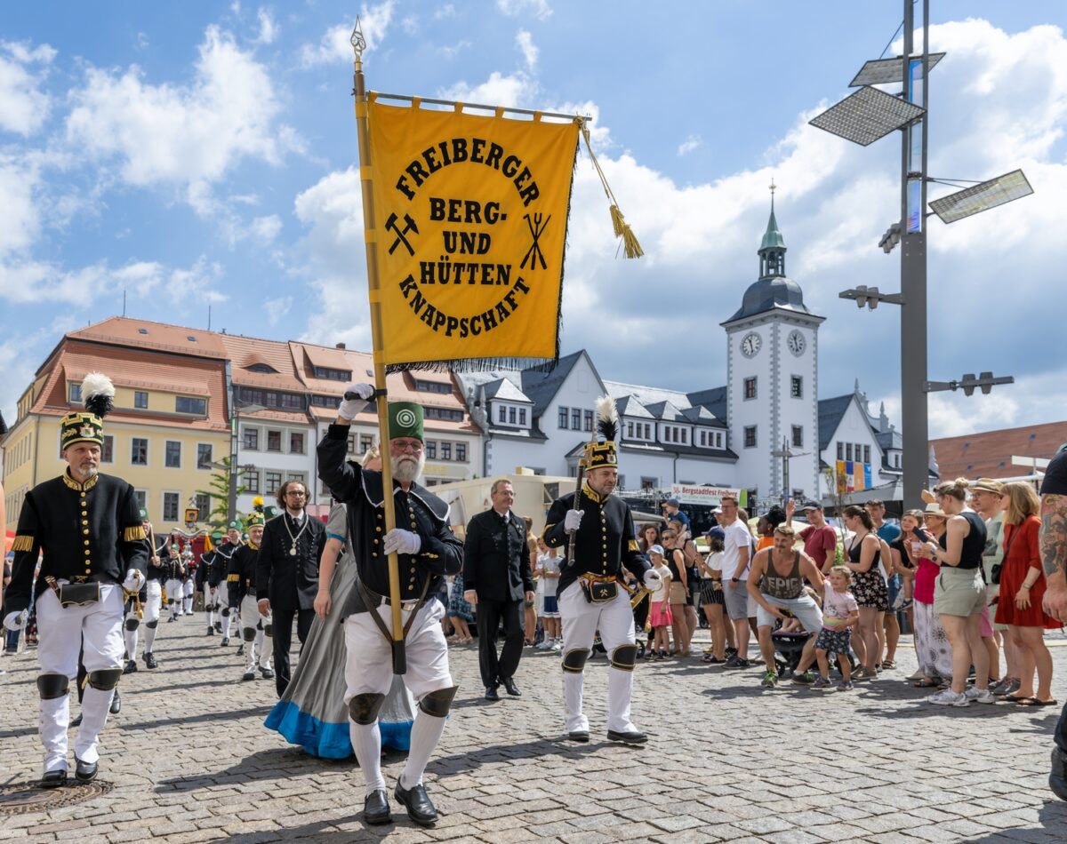 Ein Umzug mit Menschen in traditionellen Bergmannsuniformen marschiert während des Bergstadtfestes über einen Stadtplatz, angeführt von einer Gruppe, die ein gelbes Banner mit der Aufschrift Freiberger Berg- und Hüttenknappschaft hält. Zuschauer schauen zu; im Hintergrund sind historische Gebäude zu sehen.