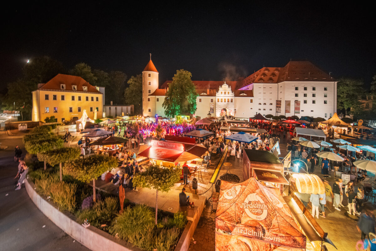 Ein lebhaftes Bergstadtfest unter freiem Himmel mit vielen Menschen, Essensständen und bunten Lichtern vor einem historischen Schloss mit Türmen und roten Dächern. Schirme und Bäume schmücken die festliche Szene.