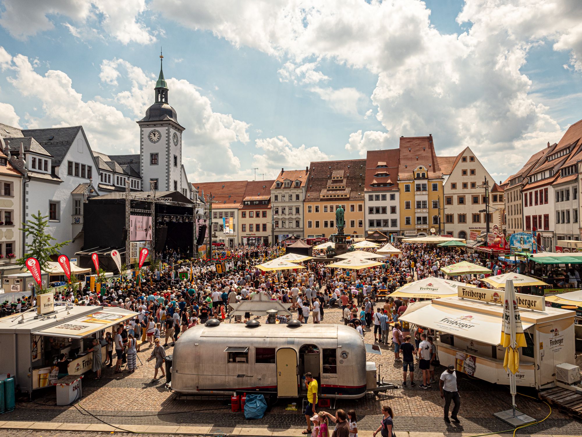 Eine große Menschenmenge füllt einen belebten Stadtplatz für das Bergstadtfest, mit Imbisswagen, Sonnenschirmen und einer Bühne in der Nähe historischer europäischer Gebäude unter einem teilweise bewölkten Himmel. Im Hintergrund erhebt sich ein weißer Uhrenturm.
