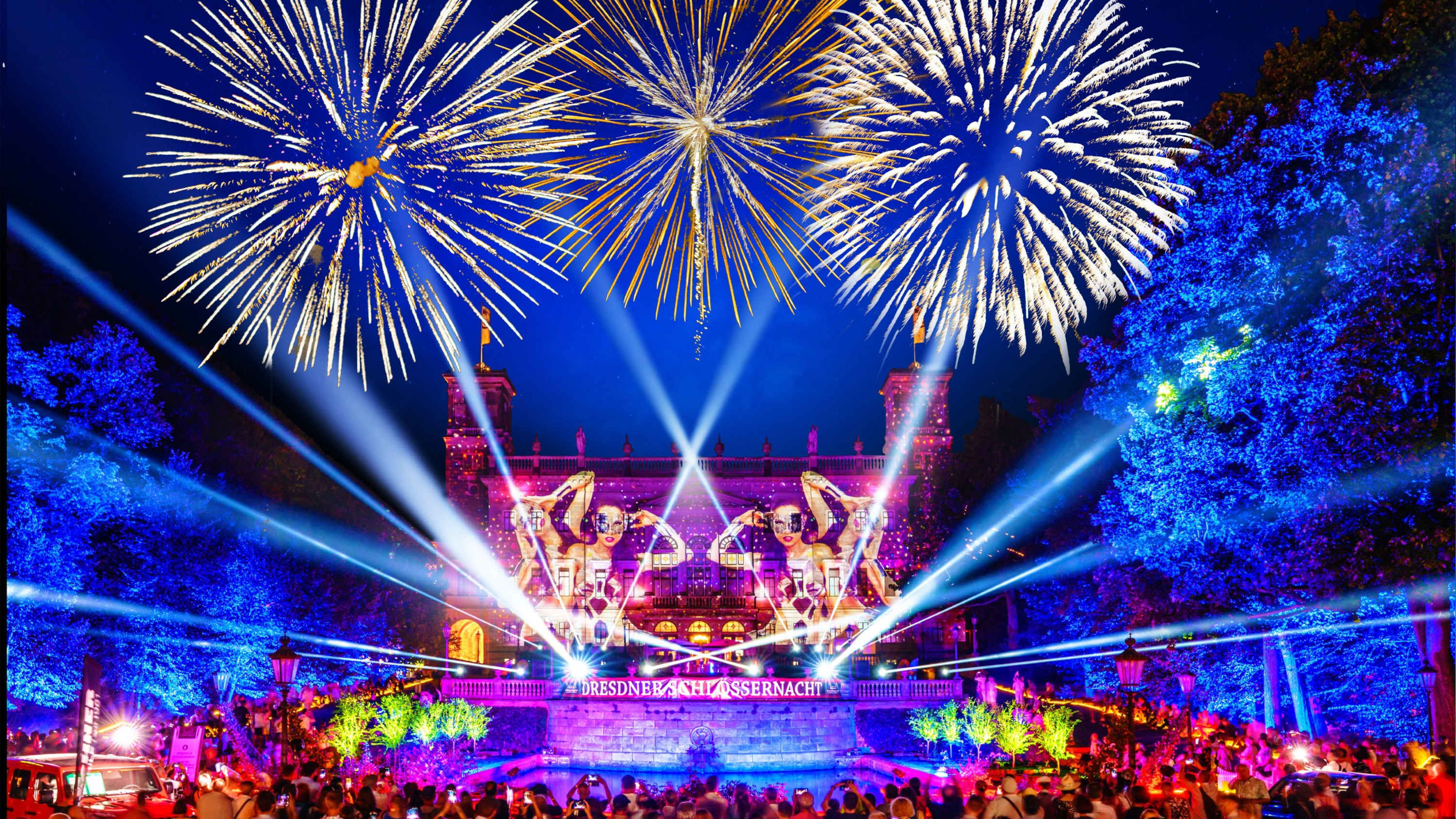 Ein lebhaftes nächtliches Fest, die Dresdner Schlössernacht, mit Feuerwerk am Himmel, bunten Lichtern, die ein großes Gebäude beleuchten, großen Statuen und einer Menschenmenge, die das Fest beobachtet.