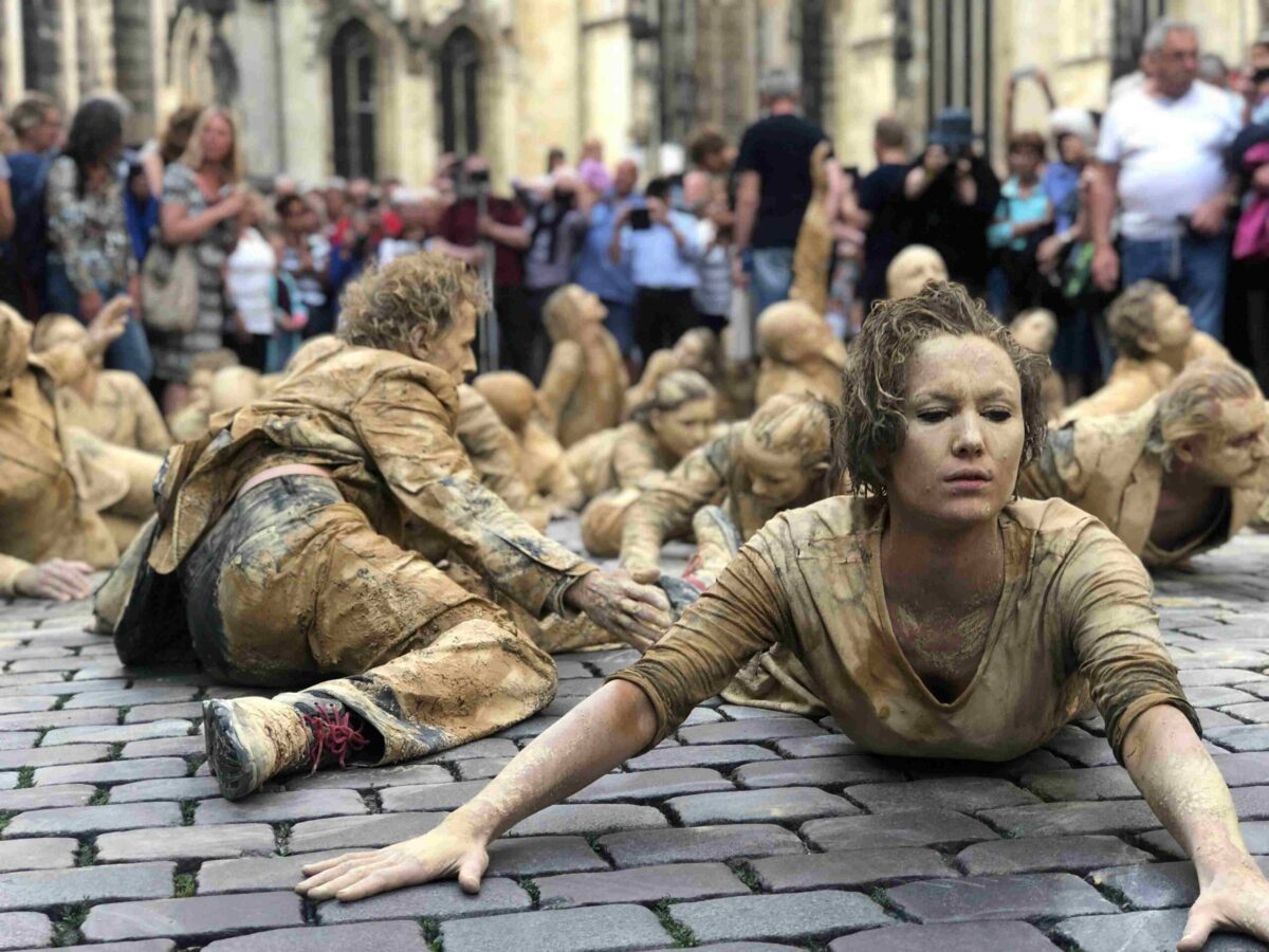 Eine Gruppe von ViaThea-Darstellern, die mit schlammfarbener Farbe bedeckt sind, kriechen und posieren dramatisch auf den Kopfsteinpflasterstraßen, während im Hintergrund eine Menschenmenge zusieht.