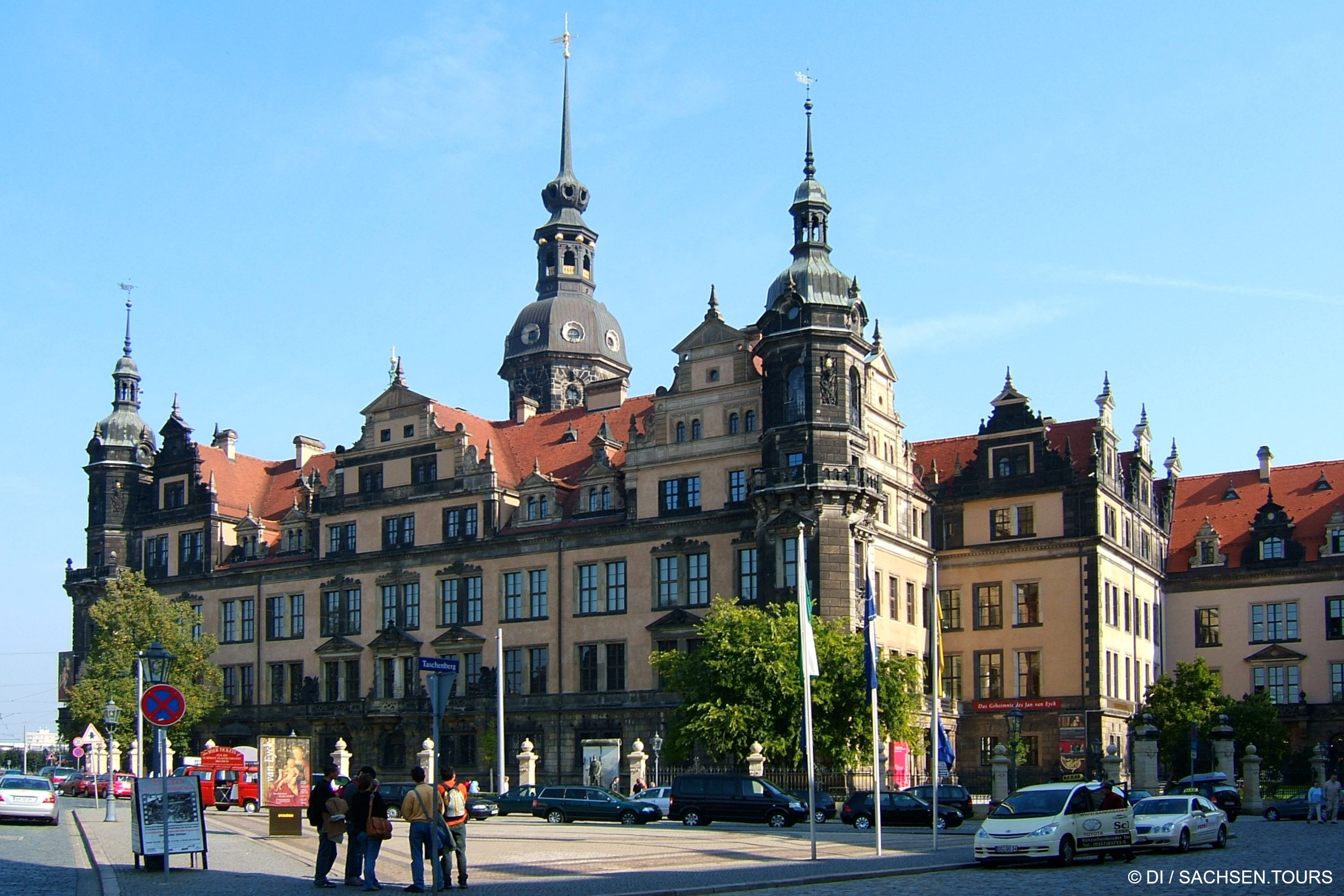 Großer verschnörkelter Renaissancepalast mit roten Ziegeldächern, Türmen und komplizierten Fassaden vor blauem Himmel. Im Vordergrund stehen mehrere Personen in der Nähe von Straßenschildern und geparkten Autos. Bäume und Laternenpfähle säumen den Bereich des Dresdner Schlosses.