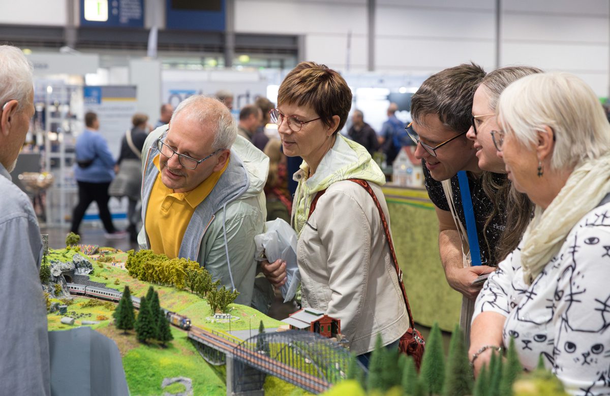 Auf der Hobbymesse betrachten fünf Erwachsene in einer Indoor-Ausstellung aufmerksam eine detailreiche Modelleisenbahnanlage; sie beugen sich vor, um die Miniaturlandschaft und die Züge zu bewundern, während andere Besucher im Hintergrund die Ausstellung erkunden.