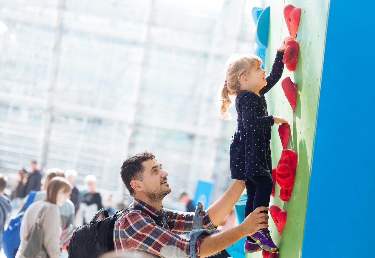Auf einer Hobbymesse hilft ein Mann einem kleinen Mädchen beim Klettern an einer Indoor-Kletterwand. Sie streckt die Hand nach einem roten Griff aus, während er sie stützt – umgeben von anderen Besuchern in einem hellen, luftigen Raum mit großen Fenstern.