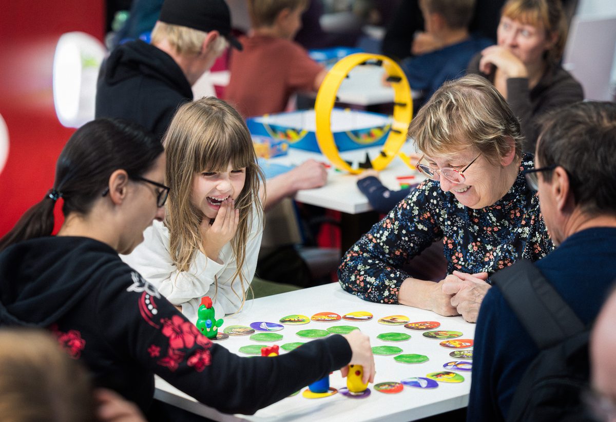 Eine Gruppe von Menschen, darunter ein Kind und ältere Erwachsene, sitzt auf der Hobbymesse um einen Tisch herum und spielt ein buntes Brettspiel; alle sind mit Begeisterung dabei und haben sichtlich Spaß. Das Mädchen lächelt und beobachtet die Spielfiguren aufmerksam, während im Hintergrund andere Gruppen ähnliche Aktivitäten genießen.