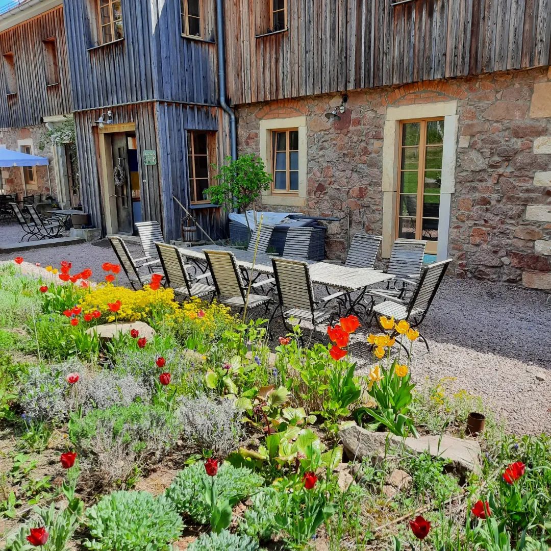 Ein langer Esstisch mit Stühlen im Freien steht auf Kies neben der rustikalen Besenwirtschaft Mariaberg aus Stein und Holz. Leuchtend rote und gelbe Blumen blühen in einem Garten im Vordergrund unter sonnigem Himmel.