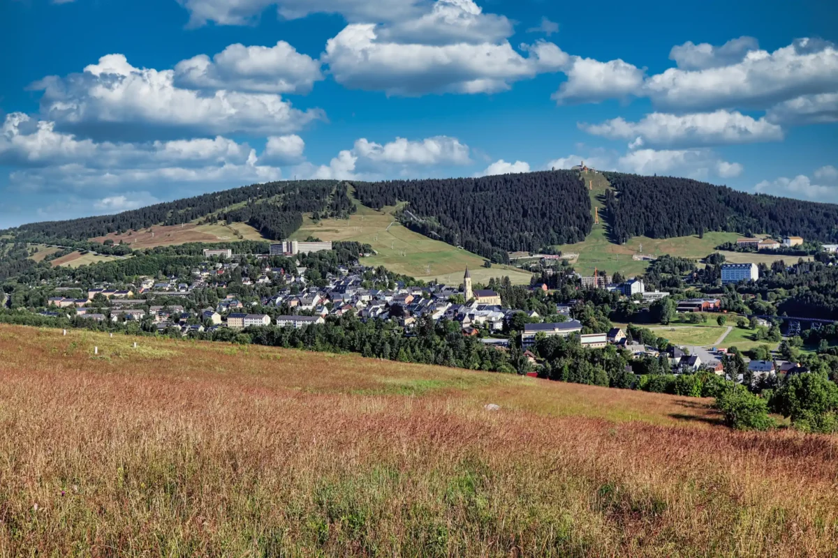 Ein Panoramablick auf eine kleine Stadt am Fuße bewaldeter Hügel, perfekt für ein Wandertag-Abenteuer, mit grasbewachsenen Feldern im Vordergrund und einem strahlend blauen Himmel mit weißen Wolken über dem Kopf.