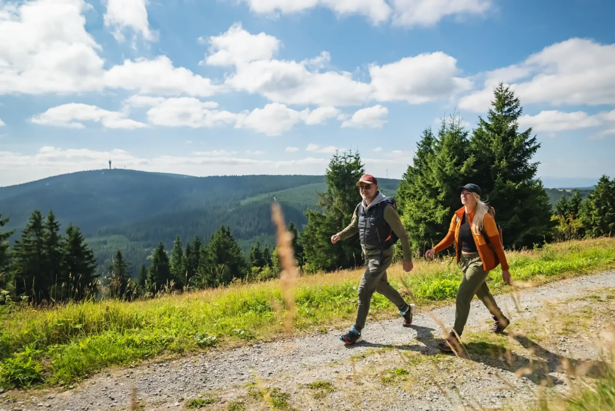 Zwei Personen genießen einen Wandertag, während sie auf einem Schotterweg wandern, der von grünem Gras und Kiefern umgeben ist, mit sanften Hügeln und einem teilweise bewölkten blauen Himmel im Hintergrund.