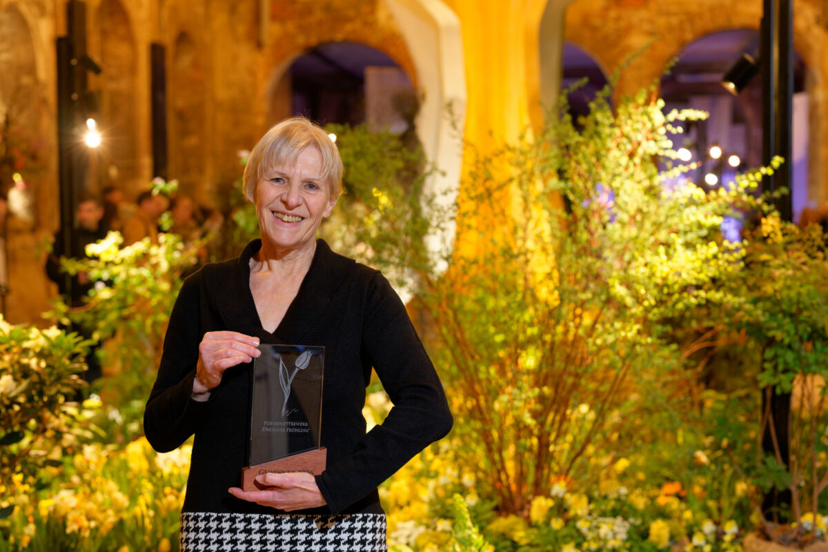 Eine lächelnde ältere Frau mit kurzen hellen Haaren hält eine Preisplakette, während sie inmitten von üppigem Grün und Blumen beim Dresdner Frühling steht, einer Indoor-Veranstaltung mit warmer Beleuchtung.