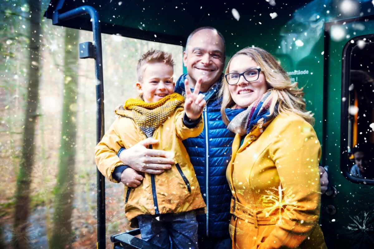 Eine lächelnde, dreiköpfige Familie in Winterkleidung steht auf einer Bimmelbahn, während um sie herum Schneeflocken fallen. Das Kind lacht, während der Lichterglanz vor der Kulisse eines winterlichen Waldes funkelt.
