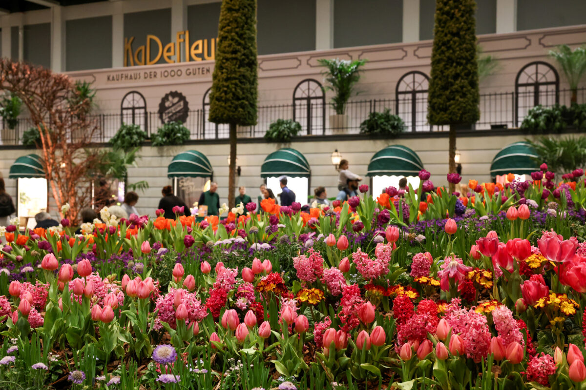 Im Vordergrund ist ein lebendiger Garten mit blühenden Tulpen und anderen farbenfrohen Blumen zu sehen, während im Hintergrund Menschen in der Nähe eines Gebäudes mit grünen Markisen und großen Fenstern stehen und gehen.