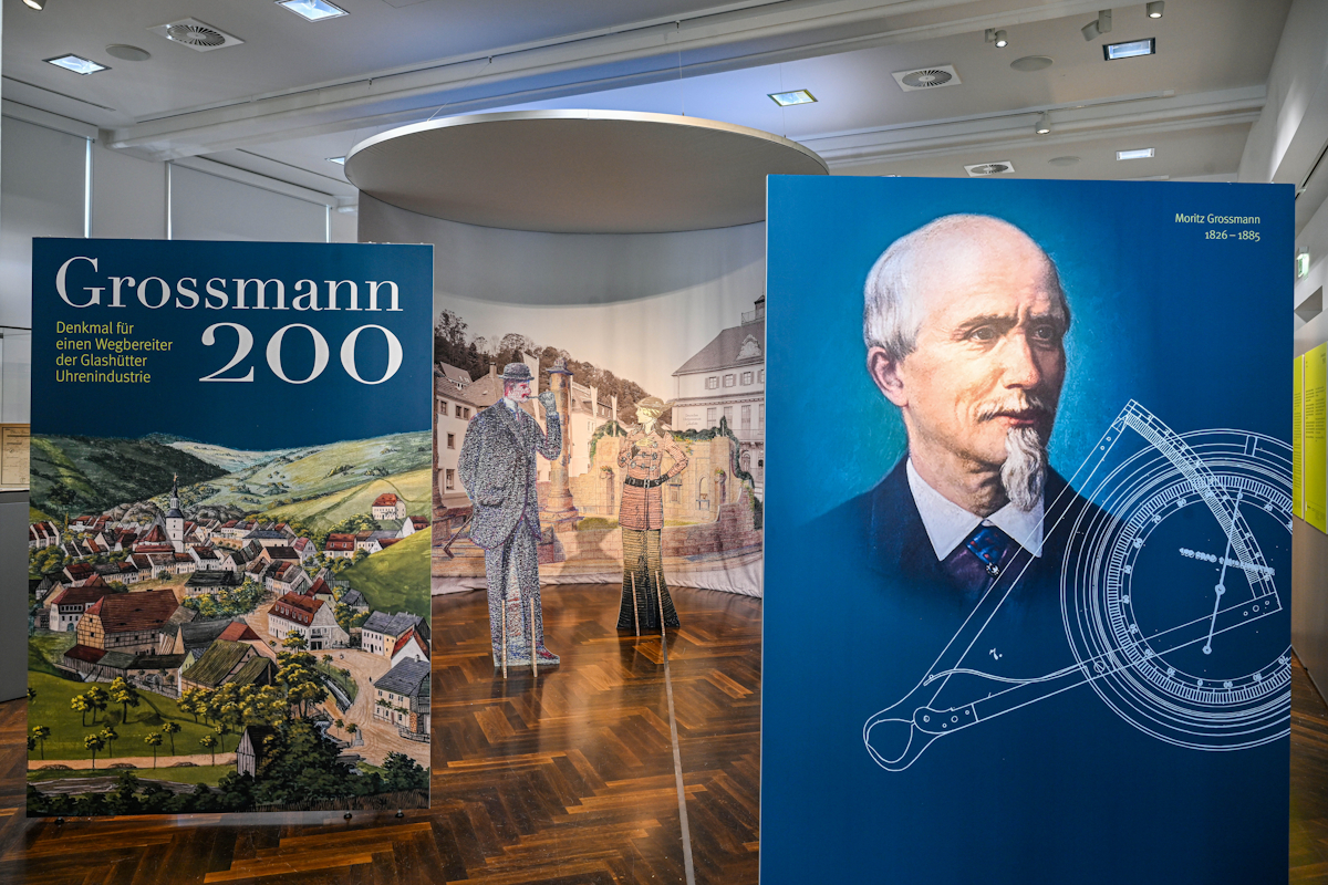 Eine Museumsausstellung mit dem Titel „Grossmann 200“, bestehend aus historischen Dorfkunstwerken, lebensgroßen Pappfiguren und einer blauen Tafel mit dem Porträt eines Mannes mit wissenschaftlichen Instrumenten, die das Vermächtnis von Moritz Grossmann durch eine öffentliche Präsentation hervorhebt.