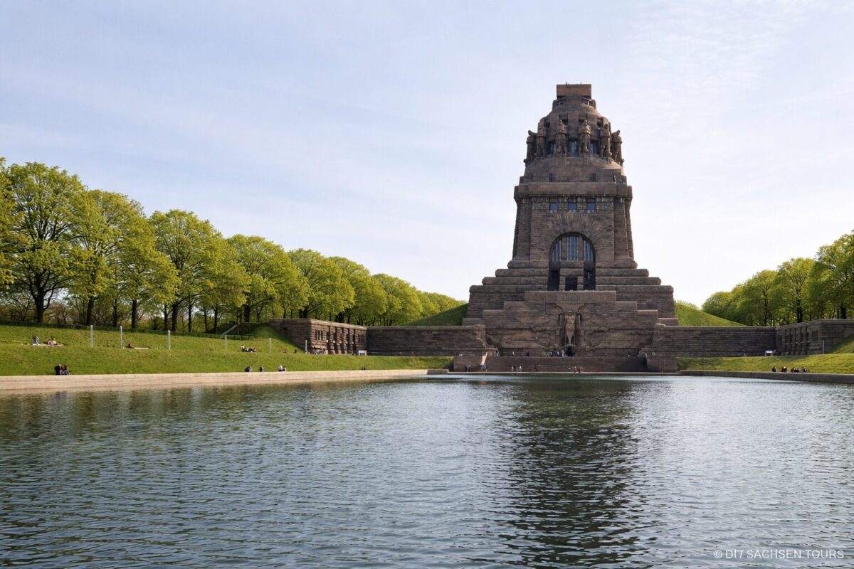 Ein großes steinernes Denkmal, das Völkerschlachtdenkmal, steht am Rande eines spiegelnden Wasserbeckens, umgeben von grünen Bäumen und Grasflächen, auf denen sich Menschen tummeln, unter einem klaren blauen Himmel.