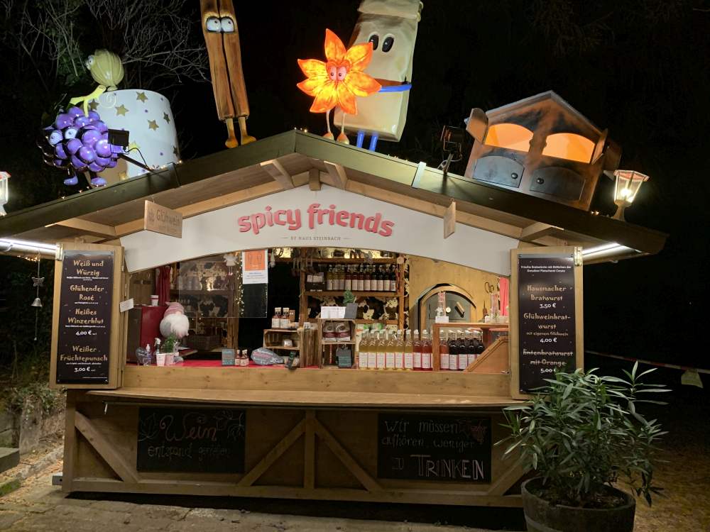 Ein hölzerner Marktstand namens Spicy Friends in Radebeul ist nachts festlich beleuchtet, mit leuchtenden Cartoon-Figuren auf dem Dach. Auf Kreidetafeln sind die Speisen aufgelistet, während Flaschen, Gläser und Glühweinzeit-Köstlichkeiten aus einem lokalen Weingut zum Stöbern einladen.
