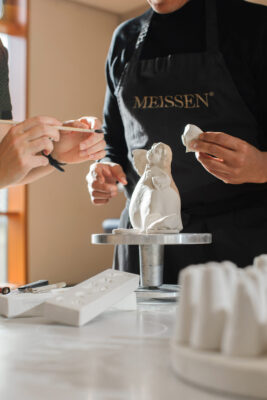 Zwei Personen arbeiten in einer Kreativwerkstatt zusammen und formen eine weiße Tierfigur aus Keramik auf einem Drehständer. Einer trägt eine schwarze Schürze mit Meissen-Aufdruck, umgeben von Werkzeugen und Formen in einem hellen, gut beleuchteten Raum.