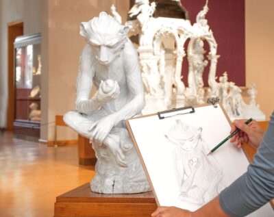 Eine Person skizziert eine detaillierte Zeichnung einer weißen Affenskulptur, die einen Apfel hält und auf einem Sockel in einer Museumsgalerie ausgestellt ist. Im Vordergrund sind die Hand und das Klemmbrett des Künstlers zu sehen.