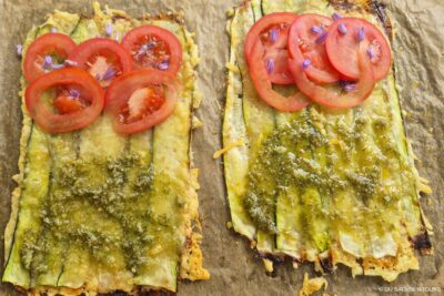 Zwei rechteckige Fladenbrote, belegt mit dünnen Zucchinischeiben; die eine Hälfte ist mit Tomatenscheiben, die andere mit grünem Pesto bestrichen. Beide sind mit lilafarbenen essbaren Blüten garniert und auf Pergamentpapier gebacken, um ein frisches, offenes Sandwich zu erhalten.