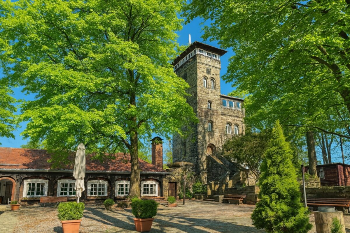 Ein historisches Steingebäude mit einem hohen Turm steht inmitten üppiger grüner Bäume. Der sonnige Innenhof des Berggasthofs Czorneboh ist mit Bänken, Topfpflanzen und einem Sonnenschirm ausgestattet und bietet eine friedliche Atmosphäre.