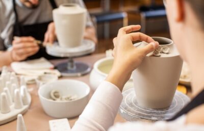 In einem Kreativworkshop bei MEISSEN dekoriert eine Person eine Vasenform Cosmopolitan mit kleinen Skulpturen, während eine andere im Hintergrund an einem Tisch im Töpferatelier arbeitet.