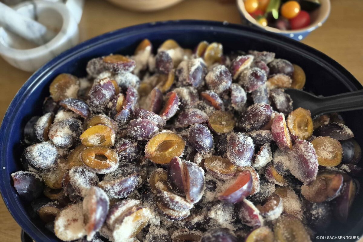 Eine blaue Schale mit halbierten und mit Zucker bestreuten Pflaumen, bereit zum Backen von Pflaumenmus. Im Hintergrund liegen Kirschtomaten in einer kleinen Schale neben einem Mörser und Stößel auf einer Holzfläche.