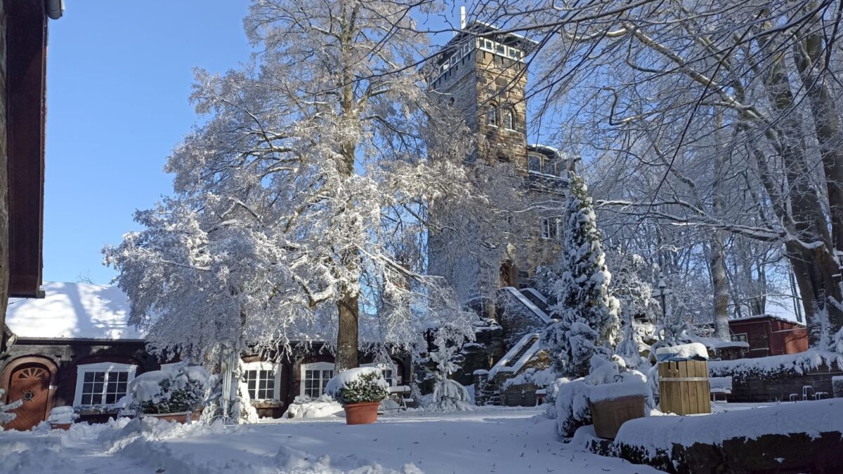 Ein steinernes Gebäude mit einem Turm steht zwischen schneebedeckten Bäumen und Topfpflanzen unter einem klaren blauen Himmel. Der Schnee bedeckt den Boden, die Dächer und die Äste des Berggasthofs Czorneboh und schafft eine friedliche Winterlandschaft.