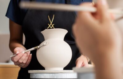 Während eines Kreativworkshops bemalt eine Person in schwarzer Schürze ein MEISSEN Vasenform Wellenspiel mit einem Pinsel, während im Vordergrund eine weitere Hand mit einem Pinsel erscheint.