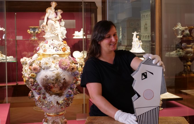 Eine Frau mit Handschuhen hantiert vorsichtig mit einer modernen, abstrakten Vase in Form eines geometrischen Gesichts, die neben einer verzierten MEISSEN-Porzellanvase in einem mit Zierkeramik gefüllten Ausstellungsraum des Museums steht.