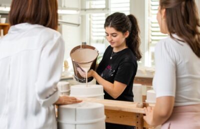 Eine Frau gießt flüssigen Ton aus einem Krug in eine Form, während zwei andere Frauen zusehen, die alle um einen Holztisch in einem hellen Atelier stehen - ein automatisch kreativer Prozess, wie ein automatisch gespeicherter Entwurf, der Gestalt annimmt.