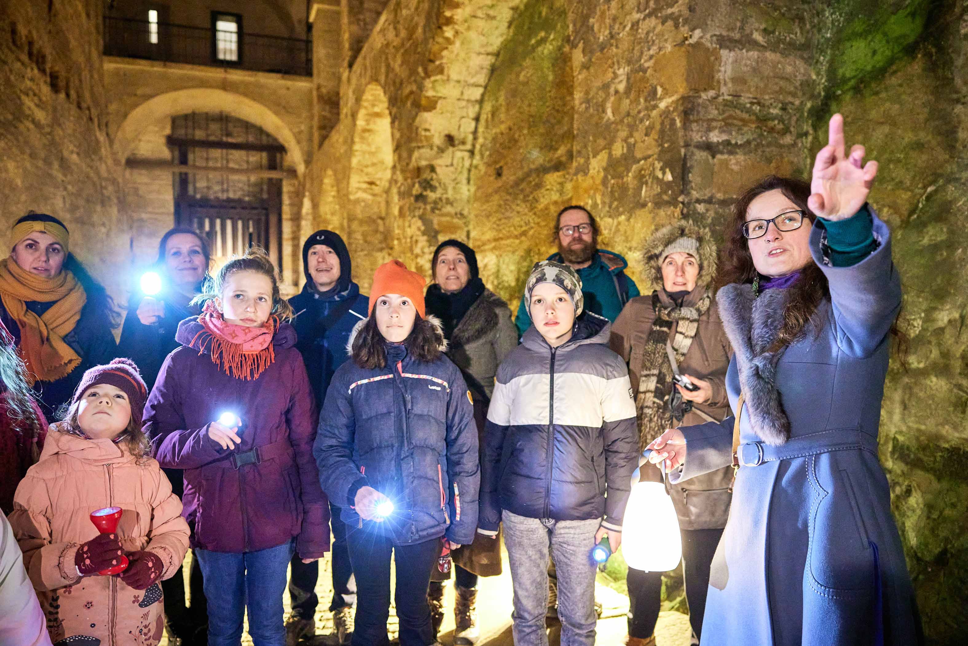 Eine Gruppe von Kindern und Erwachsenen, einige mit Taschenlampen in der Hand, hört einer Frau zu, die mit Gesten eine Taschenlampenführung in den alten Steinmauern der Festung Königstein durchführt, die von Bögen und schummrigem Licht beleuchtet werden.