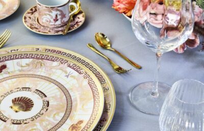 Eine elegante Tischdekoration mit exquisiter Tischkultur, bestehend aus einem gold- und rosafarbenen verschnörkelten MEISSEN-Teller, goldenem Besteck, einem Weinglas, einer Teetasse mit passender Untertasse und rosafarbenen Blumen auf einem hellgrauen Tischtuch.