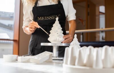 Eine Person in einer schwarzen Meißner Schürze bemalt einen weißen Keramikweihnachtsbaum, umgeben von Formen und Werkzeugen auf dem Tisch, in einem hellen, modernen Arbeitsbereich - ein automatisch gespeicherter Entwurf von Kreativität in Aktion.