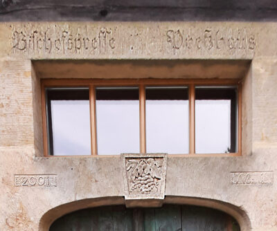 Eine Steinfassade mit einem rechteckigen Fenster mit Holzrahmen über einer Tür ist mit dekorativen Schnitzereien verziert, darunter ein Schild mit einem Löwen und den Zahlen "2901" und "1845" - eine reizvolle Station auf jedem moderierten Weinspaziergang.