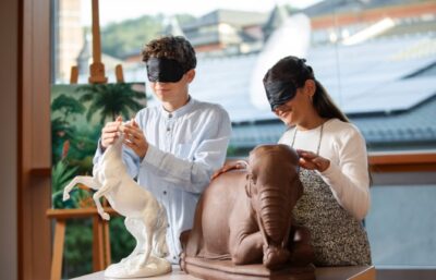 Zwei Personen mit Augenbinden berühren Tierskulpturen - ein sich aufbäumendes weißes Pferd und einen liegenden braunen Elefanten - in einem hellen Raum mit großen Fenstern und Kunstwerken im Hintergrund.