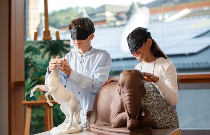 Zwei Personen mit Augenbinden berühren Tierskulpturen - ein sich aufbäumendes weißes Pferd und einen liegenden braunen Elefanten - in einem hellen Raum mit großen Fenstern und Kunstwerken im Hintergrund.