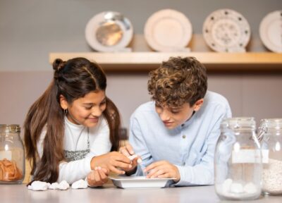 Zwei Kinder beugen sich über einen Tisch, lächeln und untersuchen Objekte, möglicherweise Steine oder Fossilien, mit Gläsern, die ähnliche Gegenstände enthalten, neben ihnen in einem hellen Raum.
