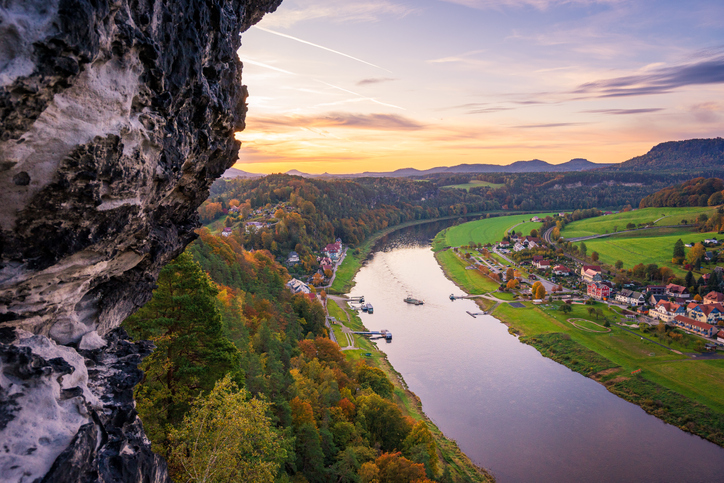 Ein malerischer Fluss schlängelt sich durch ein üppiges Tal mit farbenfrohen Herbstbäumen, kleinen Häusern und grünen Feldern - perfekt für Trailrunning - und bietet bei Sonnenuntergang einen Blick von einer felsigen Klippe unter einem Himmel mit Wolkenfetzen.