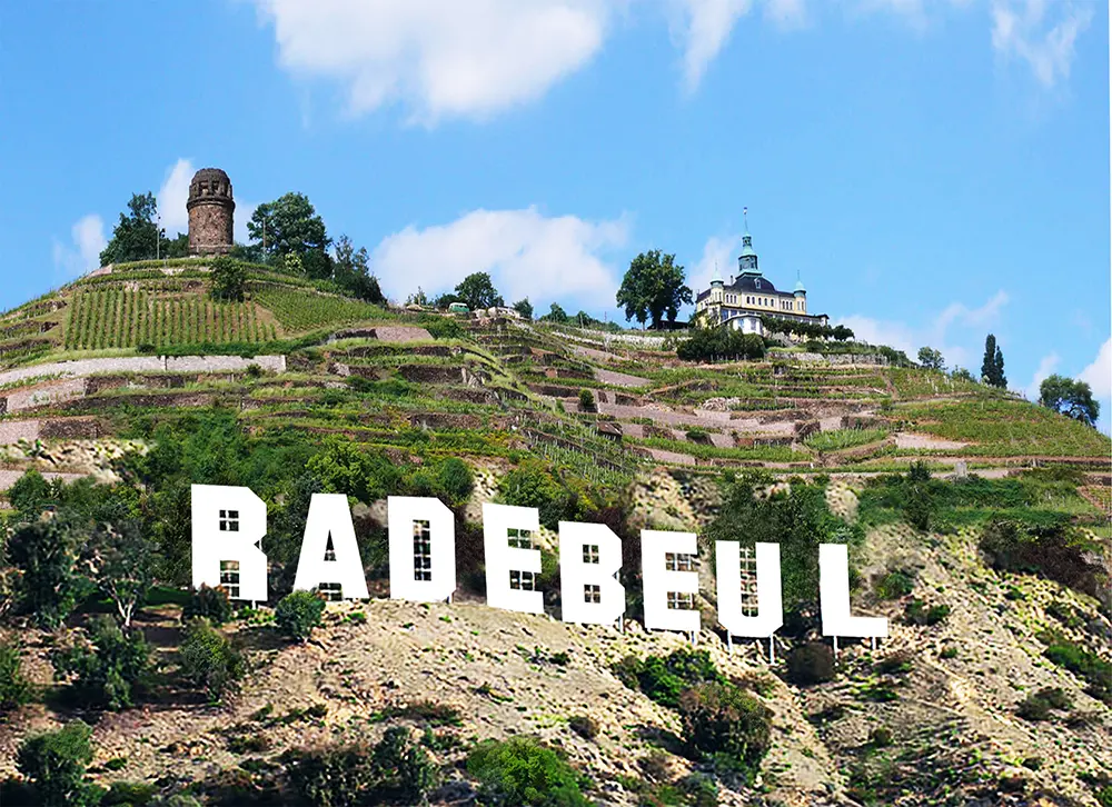 Auf einer Anhöhe mit terrassierten Weinbergen steht in großen weißen Buchstaben RADEBEUL, ähnlich dem Hollywood-Schild. In der Nähe des Gipfels steht das Haus Steinbach neben einem alten Turm, beides Wahrzeichen der landschaftlich reizvollen Weinberg KulTour unter einem blauen, wolkenverhangenen Himmel.