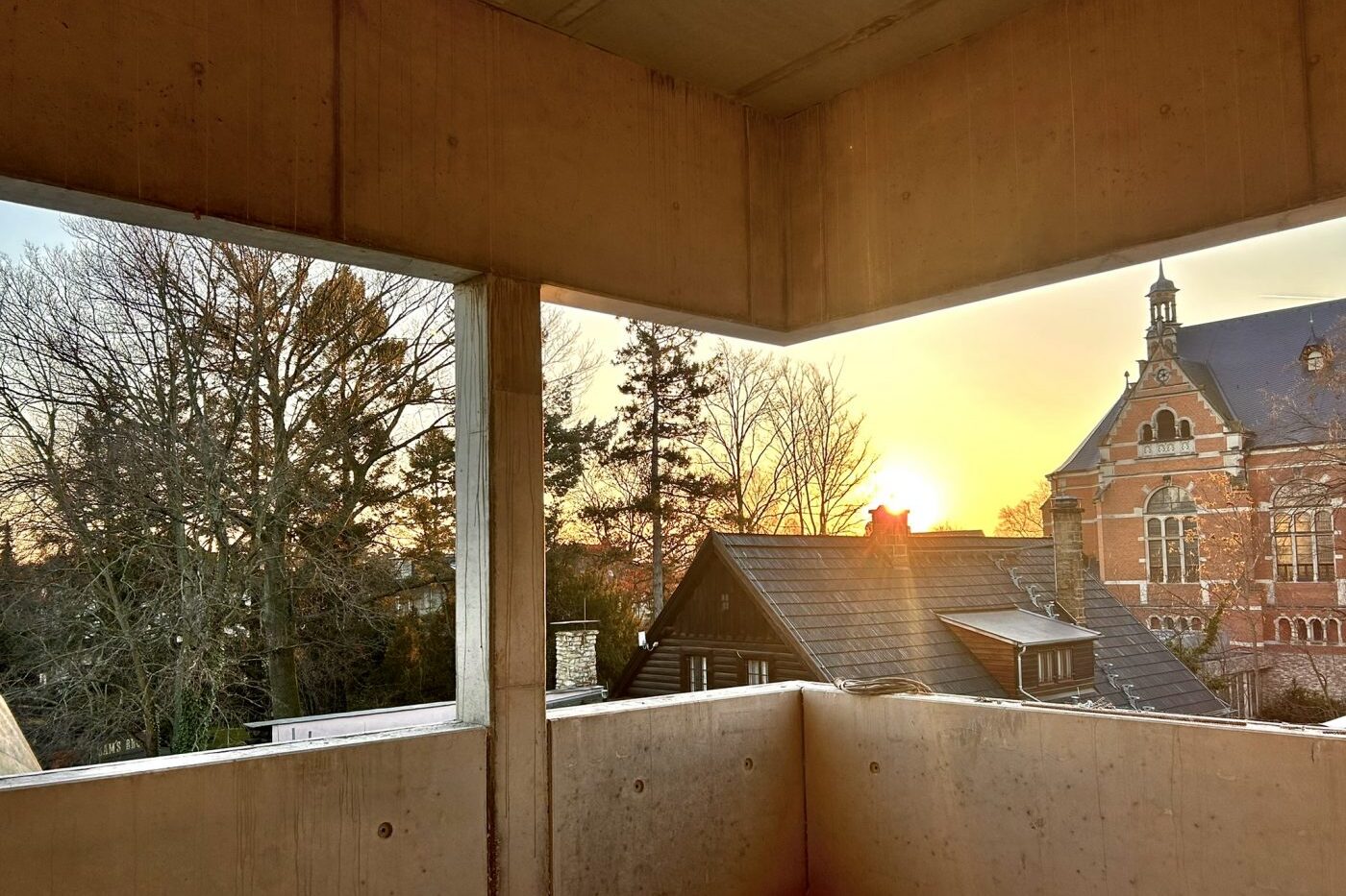 Blick auf einen Sonnenuntergang durch die offenen Betonwände eines unvollendeten Gebäudes in Radebeul, mit Blick auf Dächer, blattlose Bäume und die kunstvolle Architektur eines alten Backsteinbaus in der Nähe des Karl-May-Museums.
