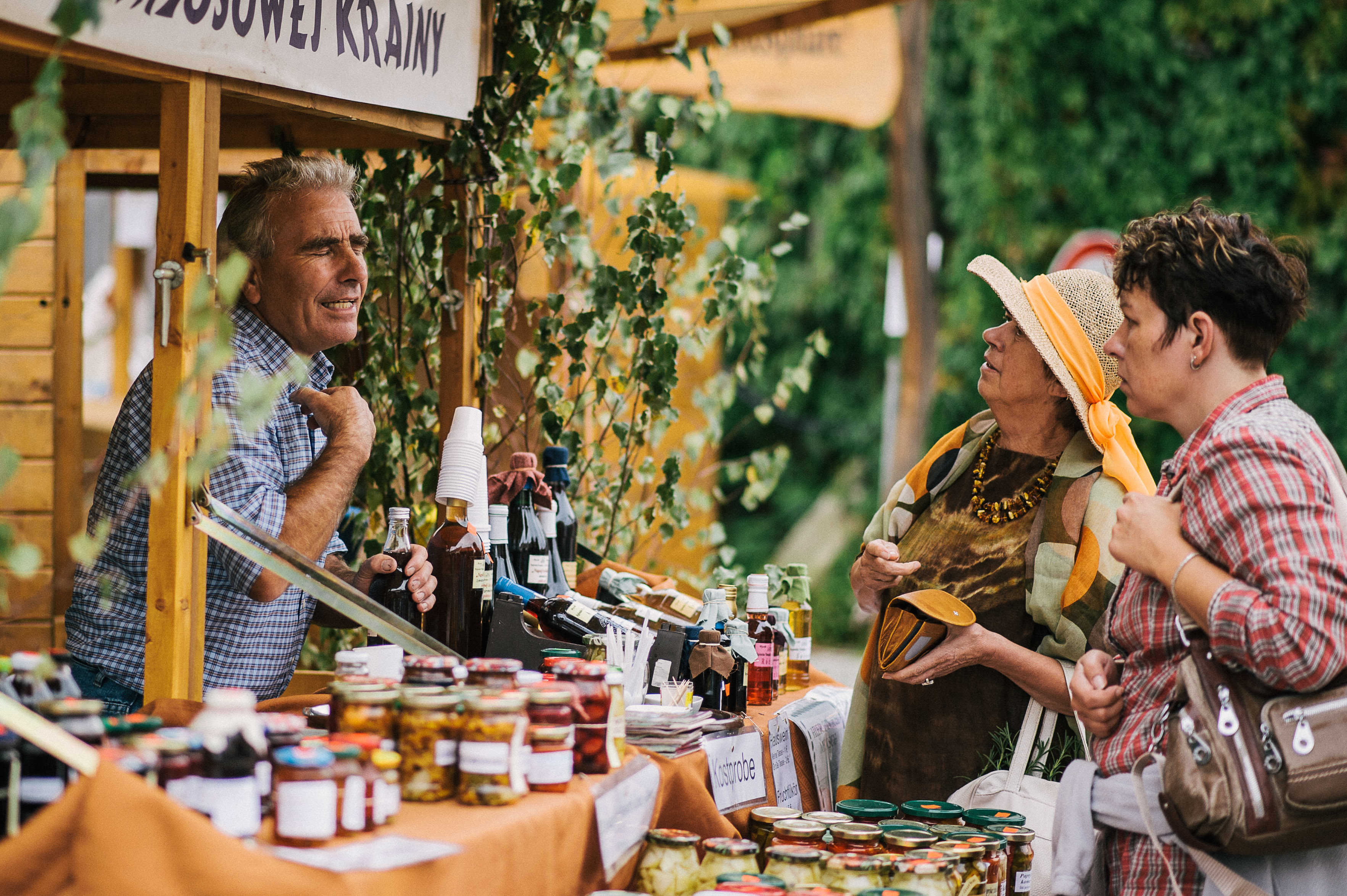 An einem Naturmarkt-Stand unter freiem Himmel lächelt ein Verkäufer und unterhält sich mit zwei Frauen, die die ausgestellten Gläser und Flaschen mit Konserven bewundern. Die Szene ist voller Grün und Holzkonstruktionen, die eine einladende Atmosphäre schaffen.