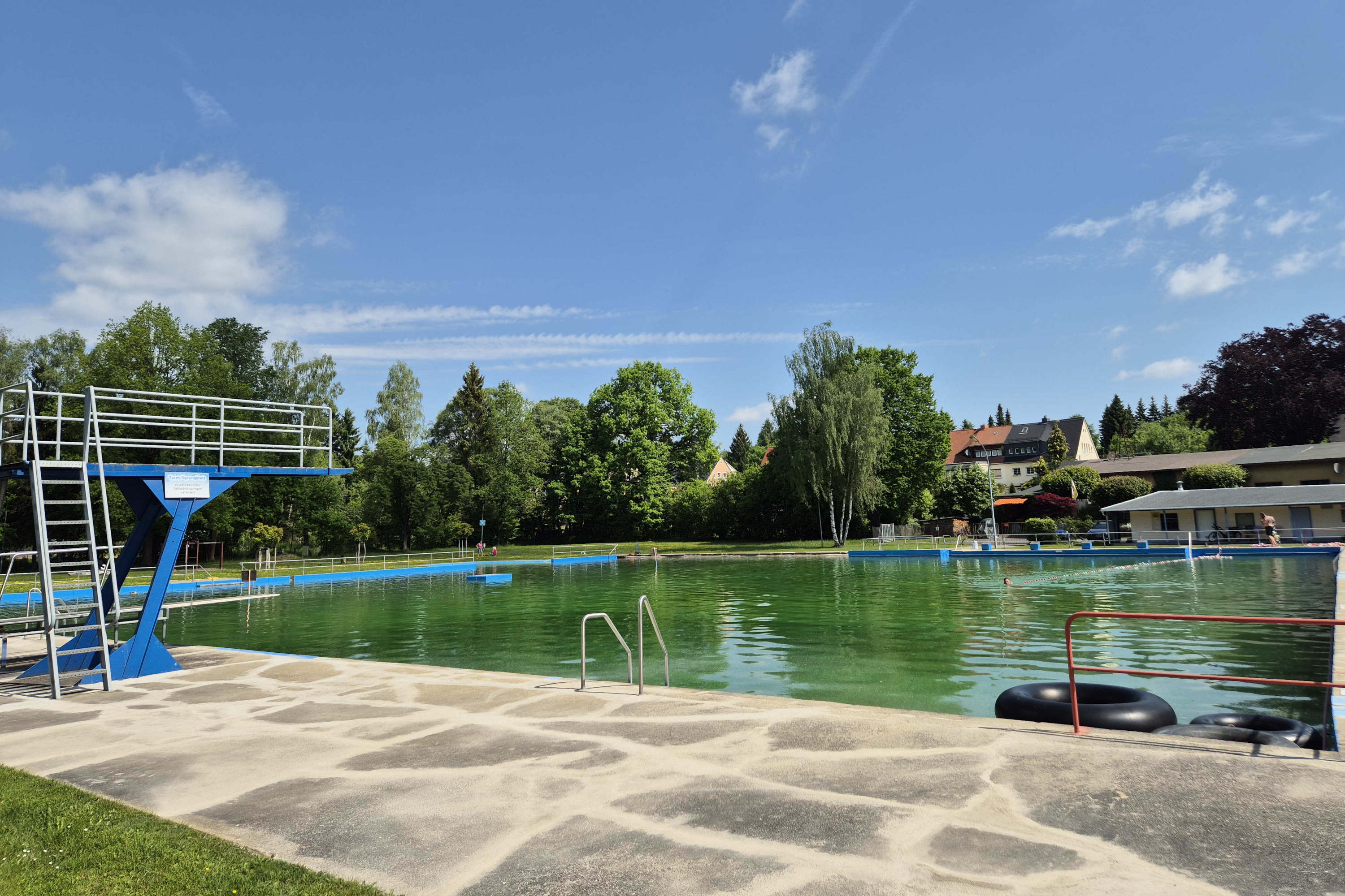 Ein Freibad mit grünlichem Wasser, links ein Sprungturm, im Hintergrund Bäume, Häuser und blauer Himmel mit Wolken. Die ruhige Badelandschaft des Oppacher Narrenbundes ist leer.