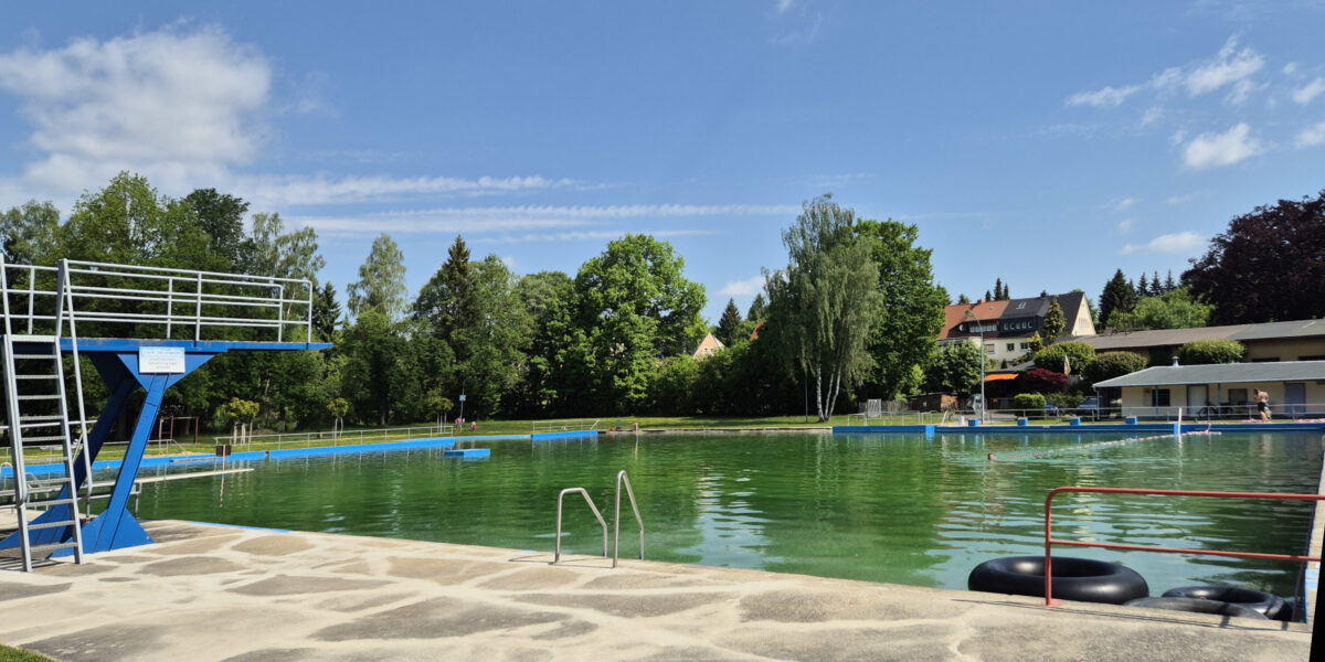 Ein öffentliches Freibad im Freibad Oppach mit grünlichem Wasser, einem blauen Sprungturm und Metallleitern. Im Hintergrund sind Bäume und Gebäude vor einem blauen Himmel mit vereinzelten Wolken zu sehen.