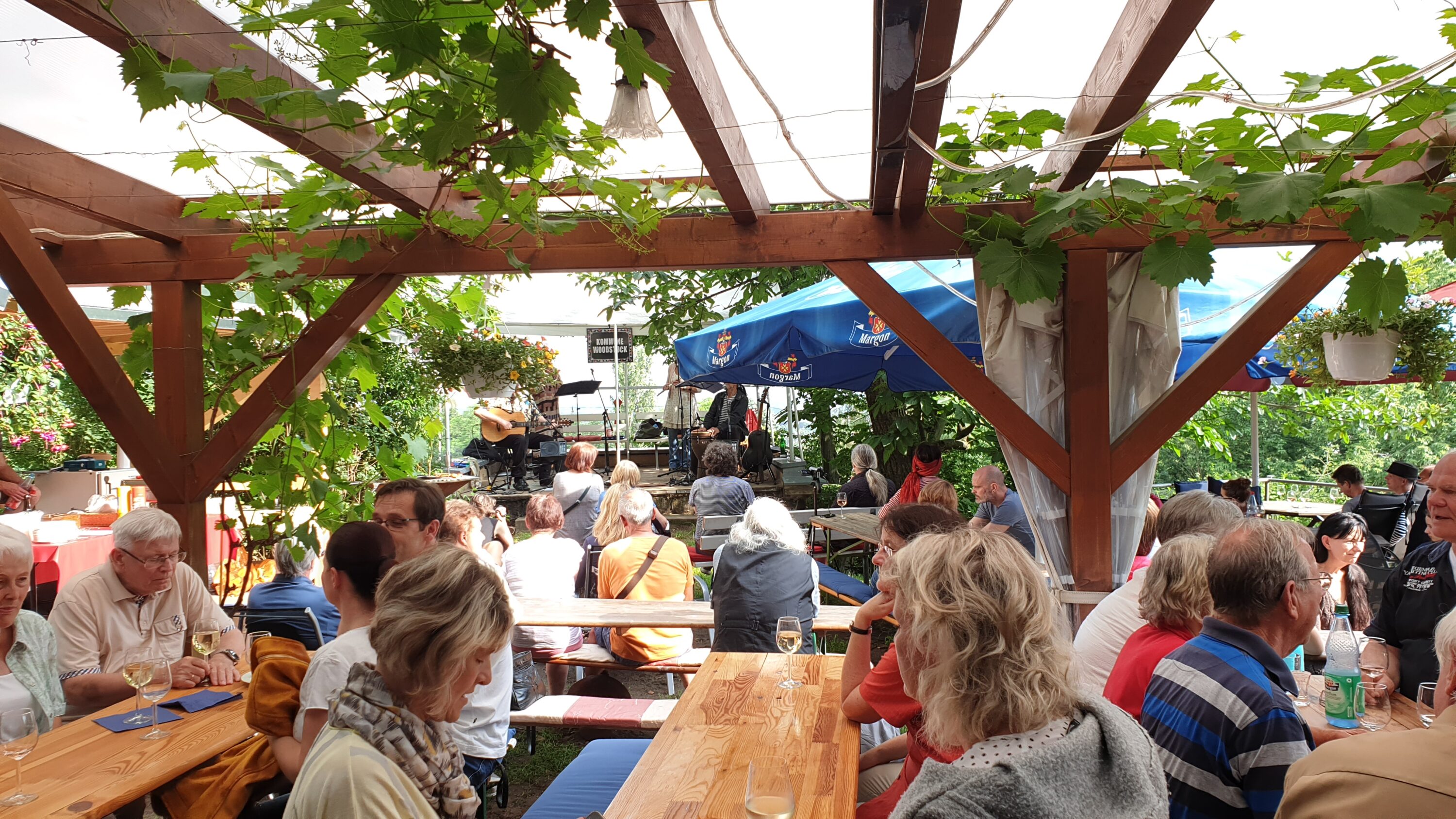 Die Gäste sitzen an Holztischen unter einer Pergola mit grünen Reben und genießen Getränke, während sie einer Band bei ihrem Auftritt im Freien zusehen. Blaue Sonnenschirme und üppiges Grün sorgen in der Gartenanlage für eine lebhafte, entspannte Atmosphäre, während der mitreißende Klang der Musik durch die Luft schwebt.