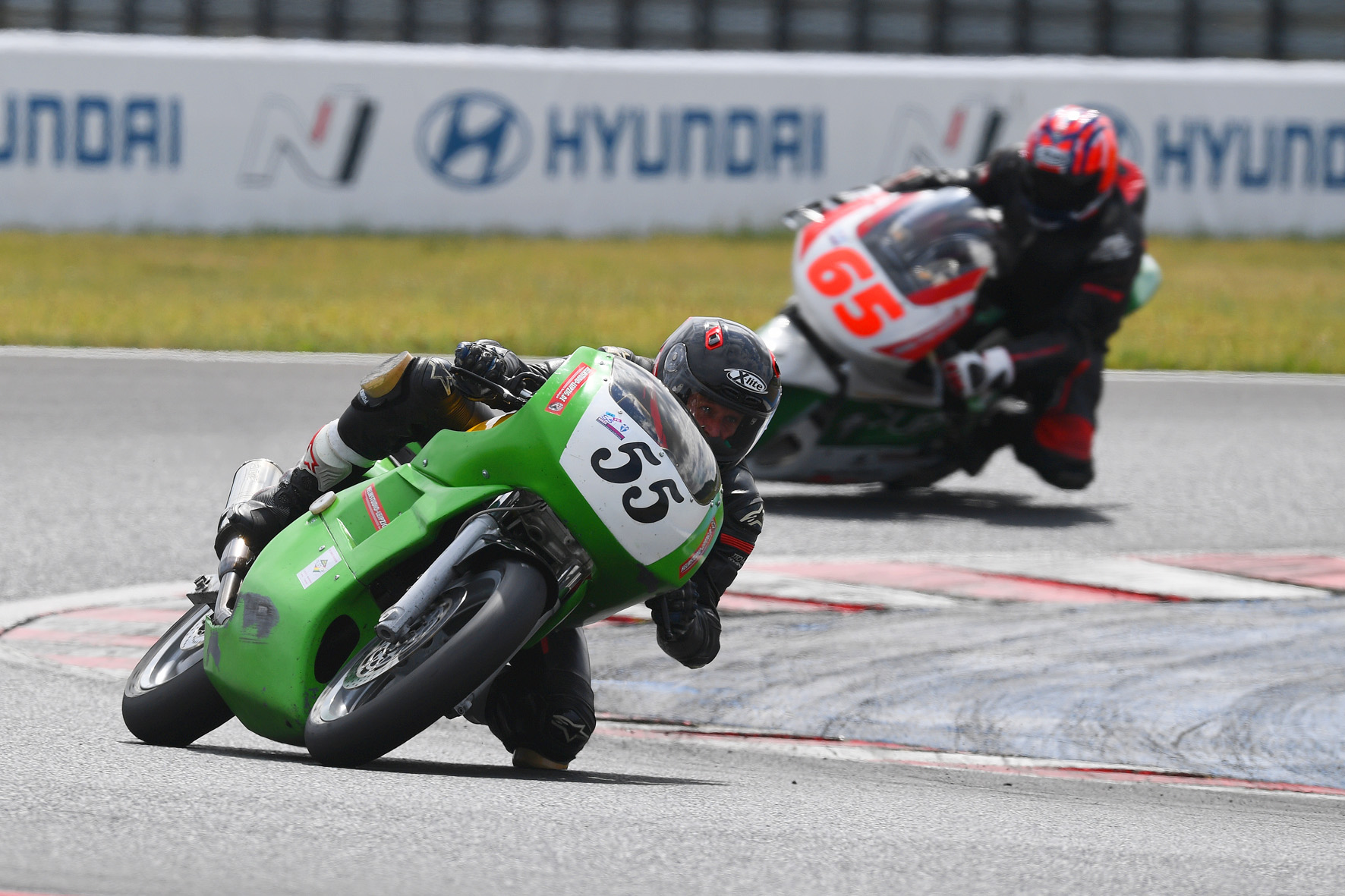 Zwei Motorradrennfahrer legen sich auf der Rennstrecke „Zschorlauer Dreieck“ in eine Kurve. Der vordere Fahrer sitzt auf einem grünen Motorrad mit der Startnummer 55, dicht gefolgt von einem Motorrad mit der Startnummer 65. Im Hintergrund ist eine Hyundai-Werbung zu sehen.