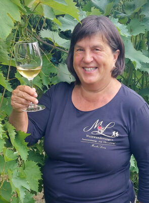 Eine lächelnde Frau hält ein Glas Weißwein in der Hand, während sie vor einer Weinrebe eine Weinwanderung genießt.