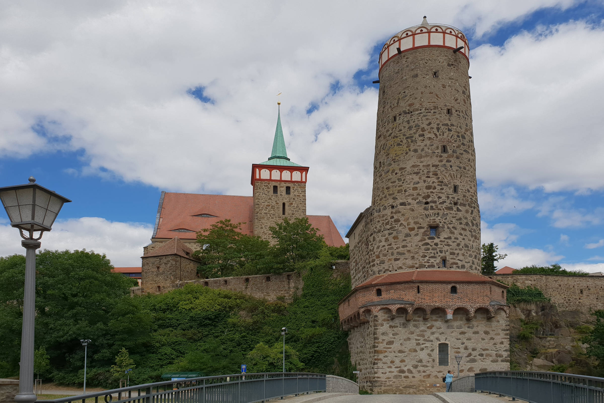 Ein hoher, runder Steinturm - Alte Wasserkunst in Bautzen - und eine Kirche mit grüner Spitze erheben sich über Bäumen und einer Steinmauer unter einem teilweise bewölkten Himmel, gesehen von einer Brücke mit einer Straßenlaterne auf der linken Seite.