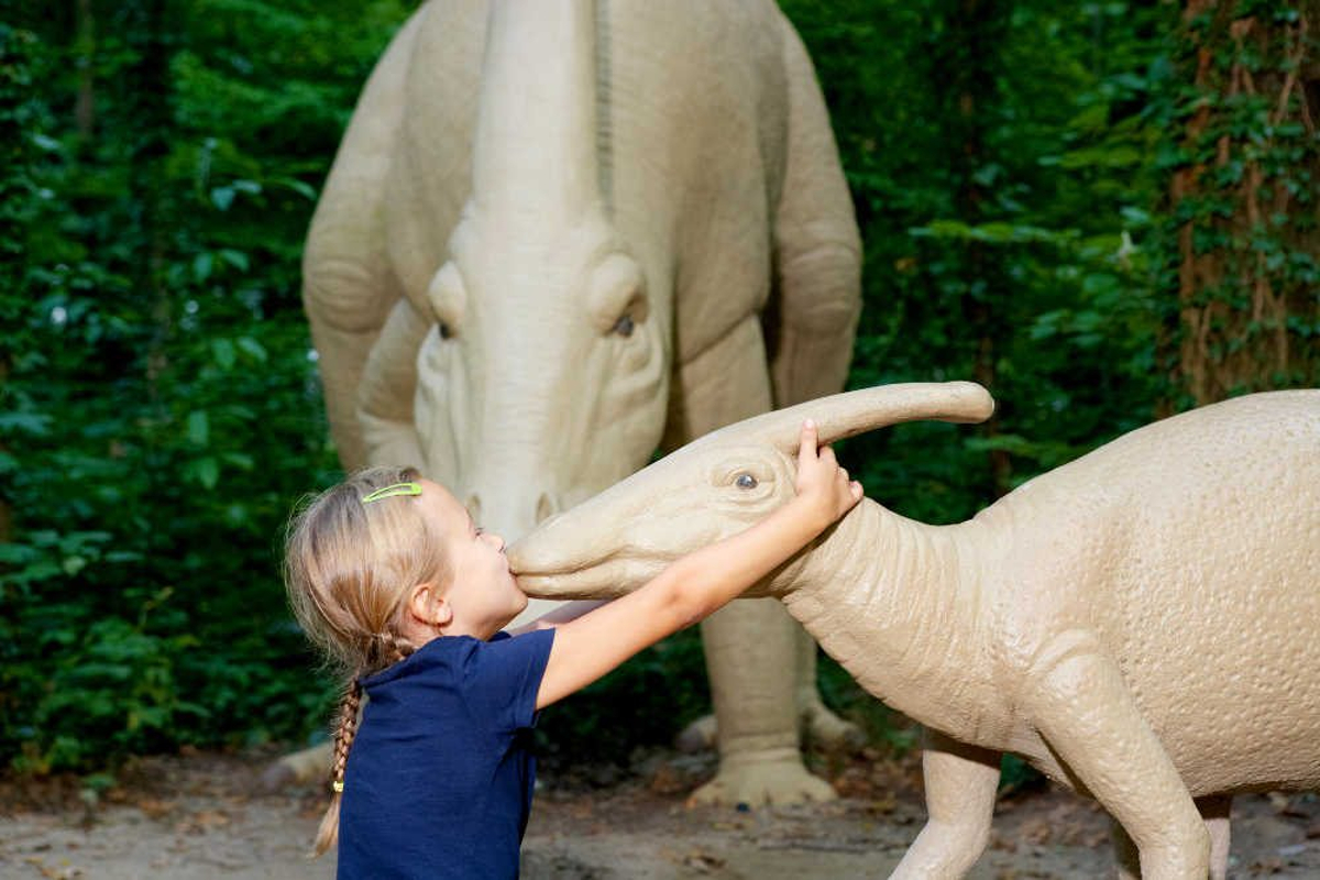 Ein junges Mädchen in einem marineblauen Hemd küsst die Schnauze einer Dinosaurier-Statue im Saurierpark Kleinwelka in Deutschland, während eine andere große Dinosaurier-Statue hinter ihr inmitten grüner Bäume im Dinosaurierpark steht.